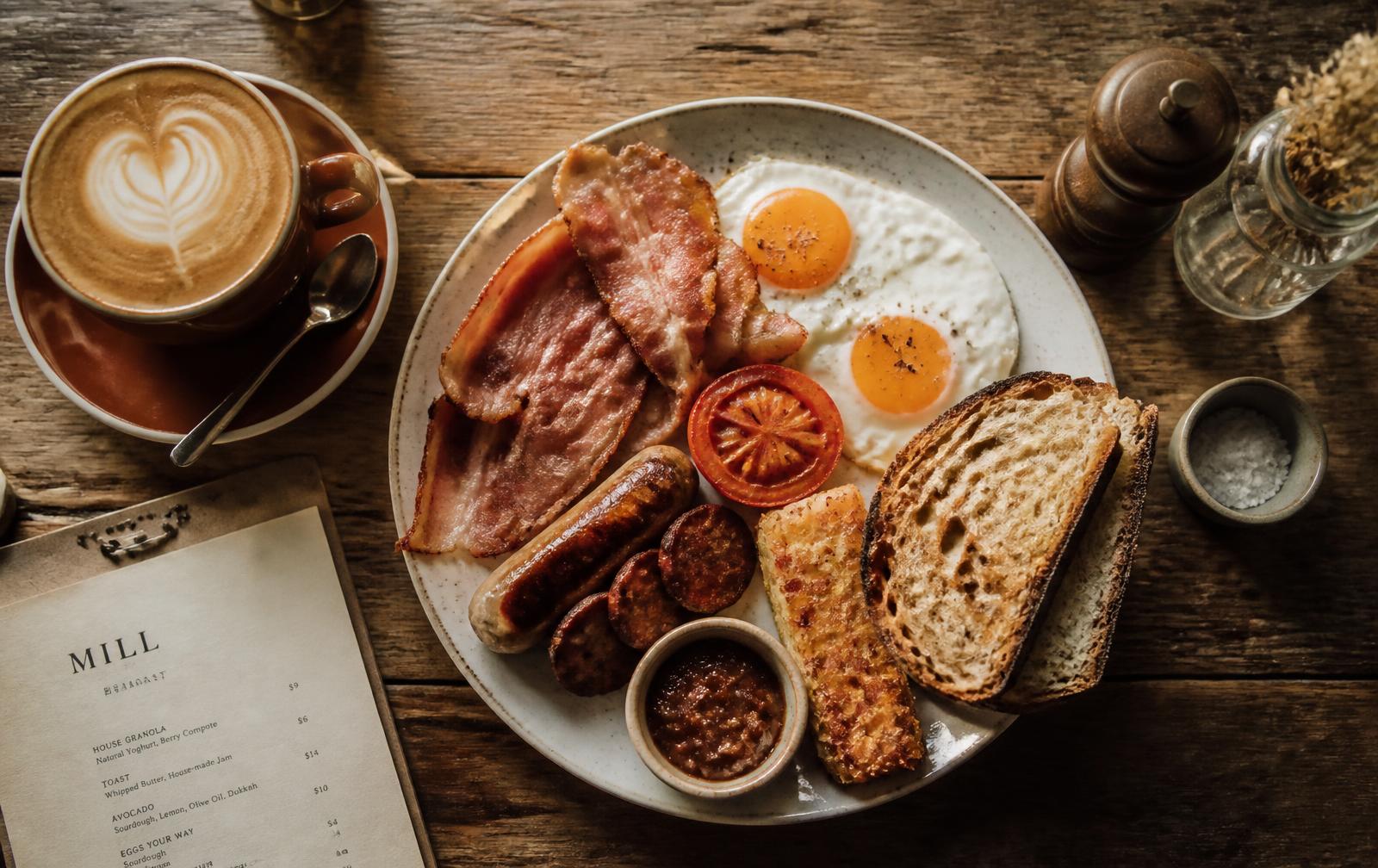 Big breakfast plate with bacon, eggs, chorizo and sourdough next to a flat white